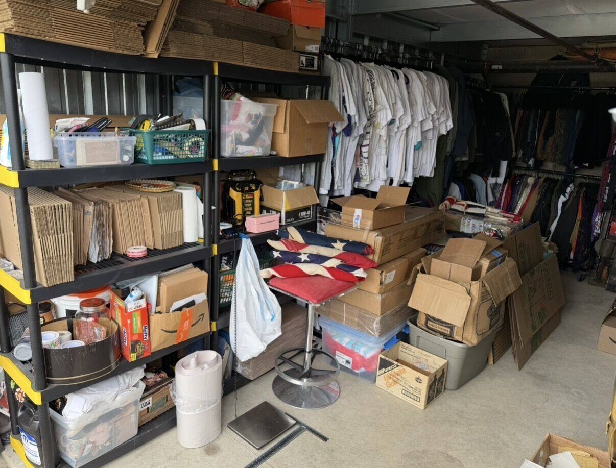 The same storage unit today: shelves of boxed inventory, shipping supplies, racks of listed clothing, and antique flags